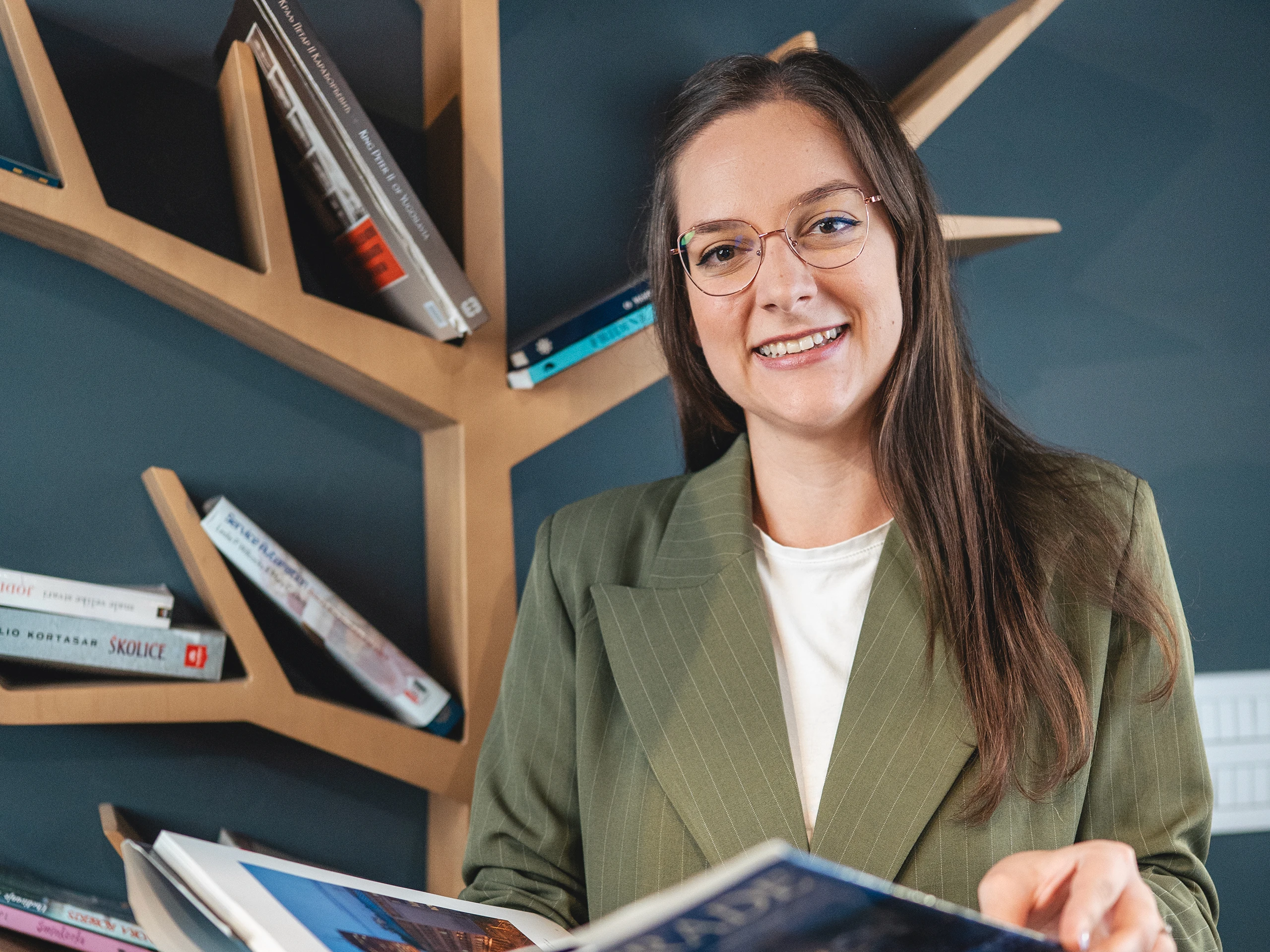 Person standing by a tree-shaped bookshelf while reading a book.