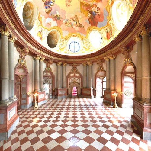 Opulent hall with a checkered marble floor, ornate columns, and a domed ceiling with detailed frescoes.
