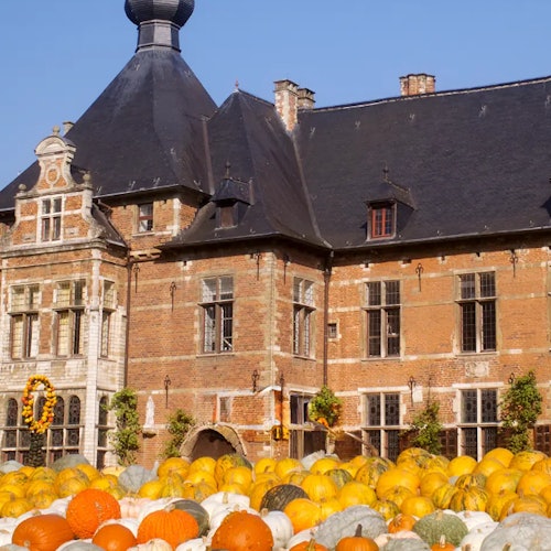 Historic brick building with a tall, dark roof, large windows, and many pumpkins of various colors in front.