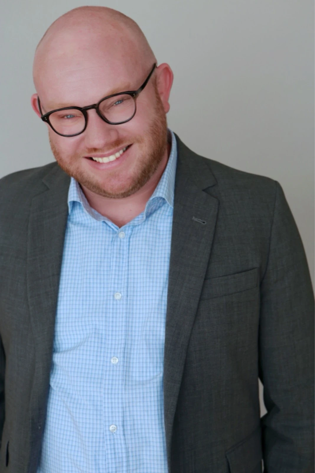 Uma pessoa usando óculos e um terno cinza-escuro sobre uma camisa xadrez azul-clara está sorrindo contra um fundo liso.