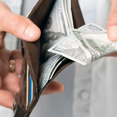 A man in casual clothes paying his bills holds a brown wallet with hundred dollar bills in his hand.