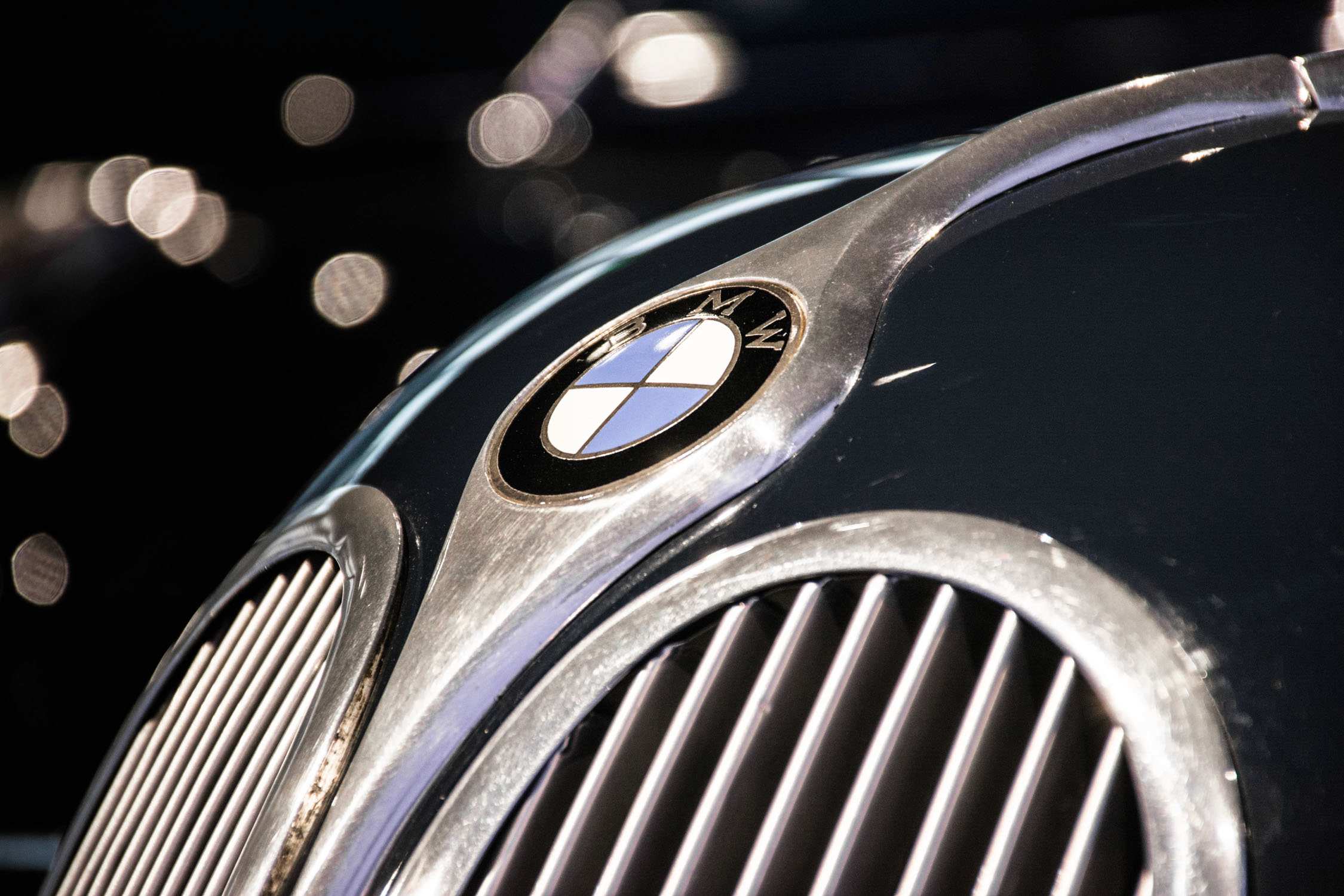 A BMW logo and chrome grille on a dark car hood with blurred lights in the background.