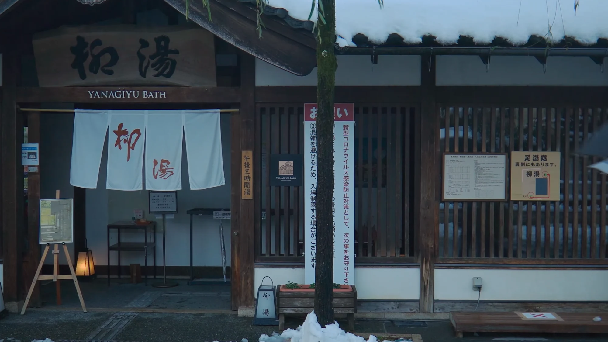 Yanagi-yu bathhouse in Kinosaki with snow on its roof and a figure under an umbrella outside.