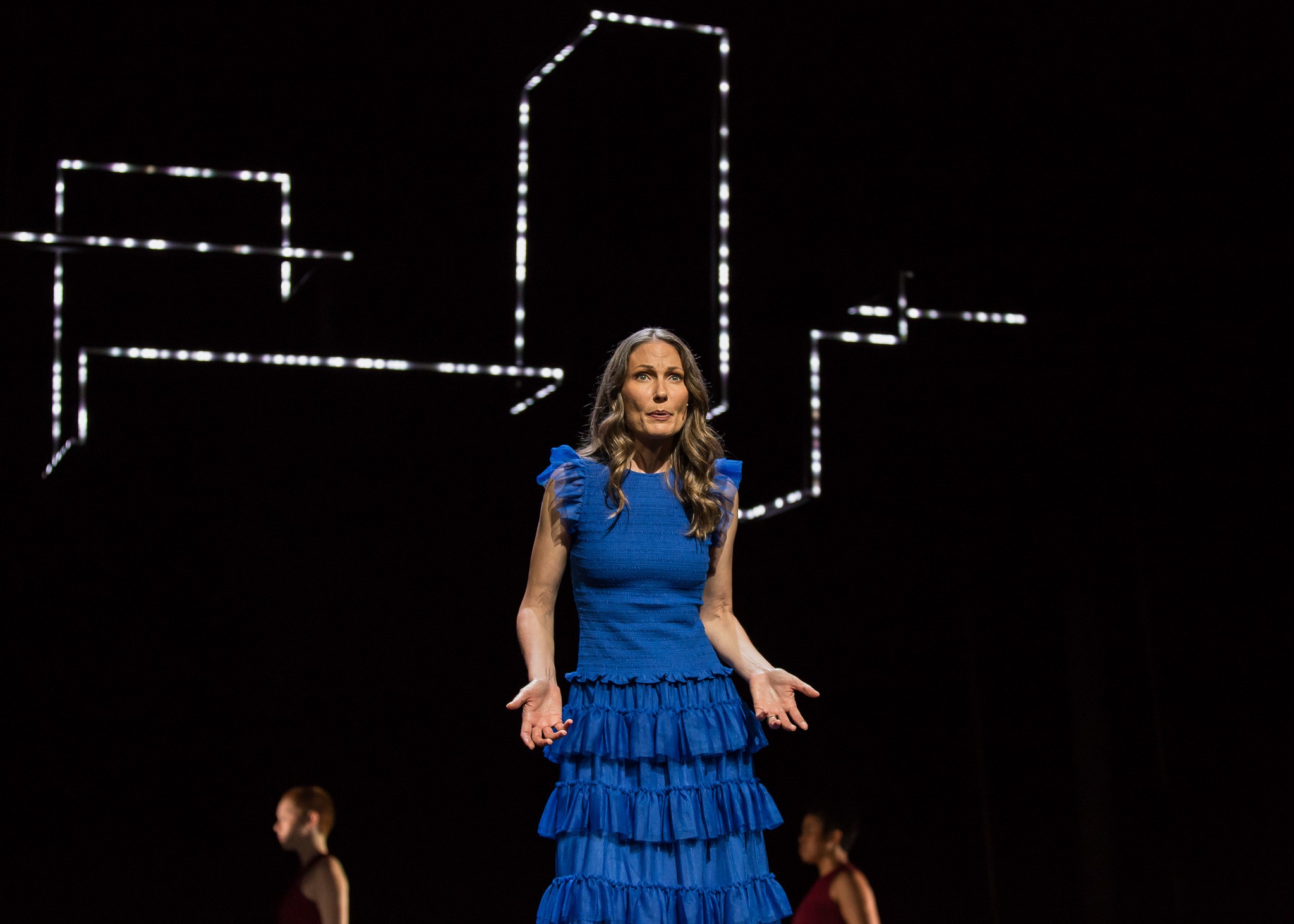 A performer in a blue dress stands on stage, illuminated against a dark background, with various lights above.