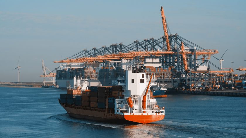 A large cargo ship carrying multicolored shipping containers sails through a bustling port with towering cranes and wind turbines in the background, highlighting industrial shipping and renewable energy.