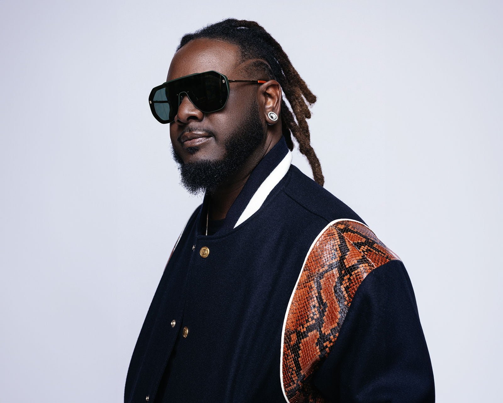 A man in stylish sunglasses and a dark jacket with brown accents poses against a plain background.