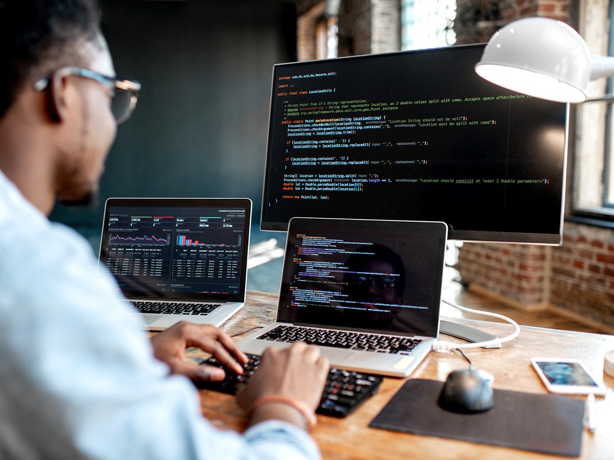 Man sitting at a desk with multiple monitors displaying lines of code, deeply focused on his tech job. The workspace is modern, with a sleek keyboard and various tech gadgets surrounding him, highlighting a busy and productive coding environment.