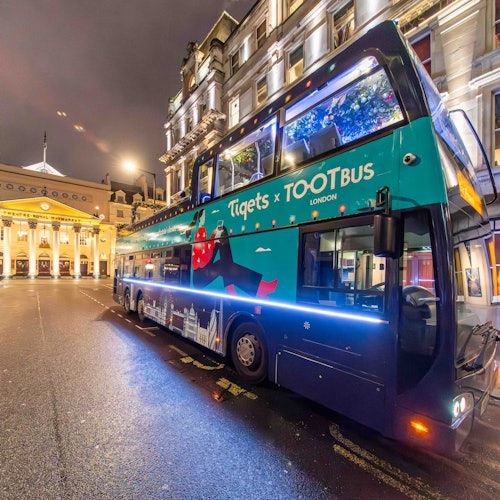 A double-decker tour bus in London with "Tiqets x TOOTbus" branding parked on a street at night near illuminated historic buildings.