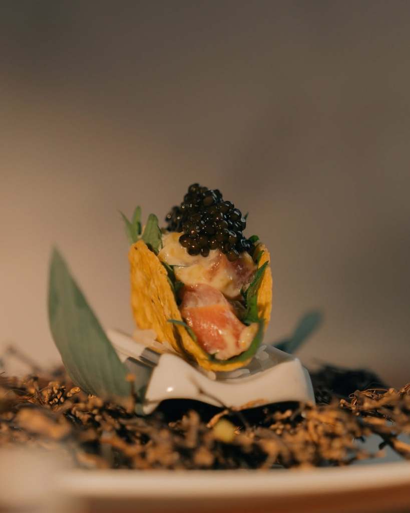 A gourmet dish featuring a crispy taco shell filled with diced seafood, creamy sauce, and topped with black caviar, presented elegantly on a decorative plate.