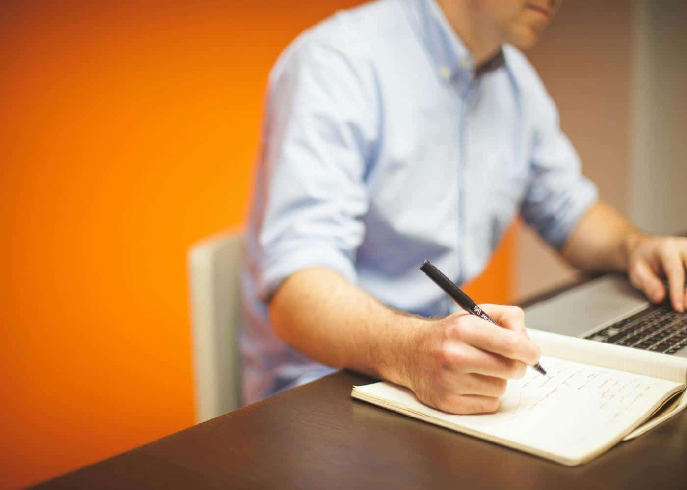 Man sitting on desk looking at laptop while taking notes on a notebook