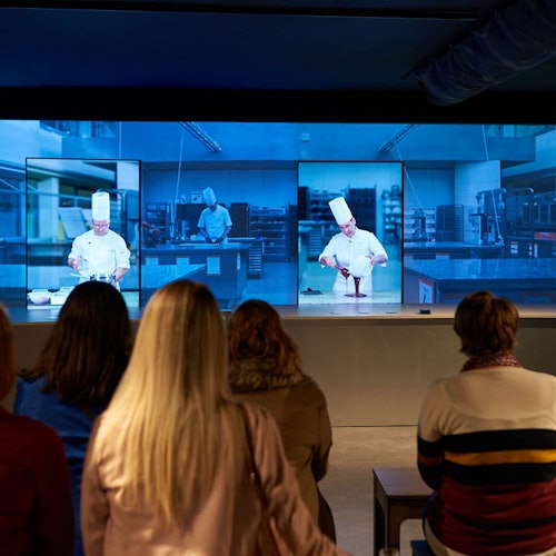 Flera personer sitter framför en stor skärm som visar tre bilder av en kock i en köksmiljö.