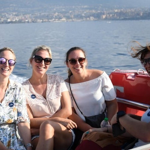 ארבע נשים, מחייכות וחובשות משקפי שמש, יושבות על סירה עם מים ונוף של העיר ברקע.