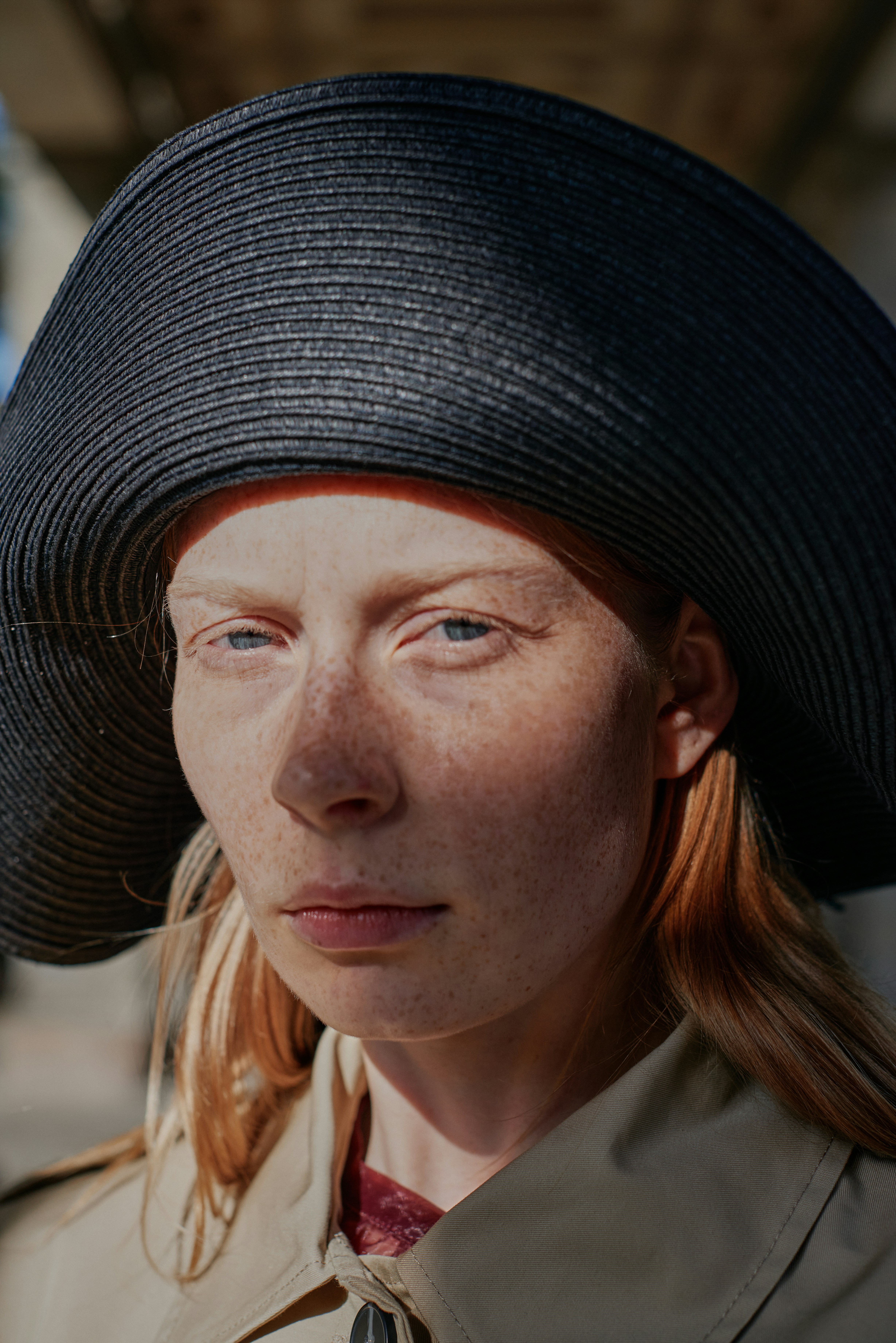 Close-up editorial portrait of a freckled redhead model wearing a wide-brim black straw hat, direct gaze, natural light