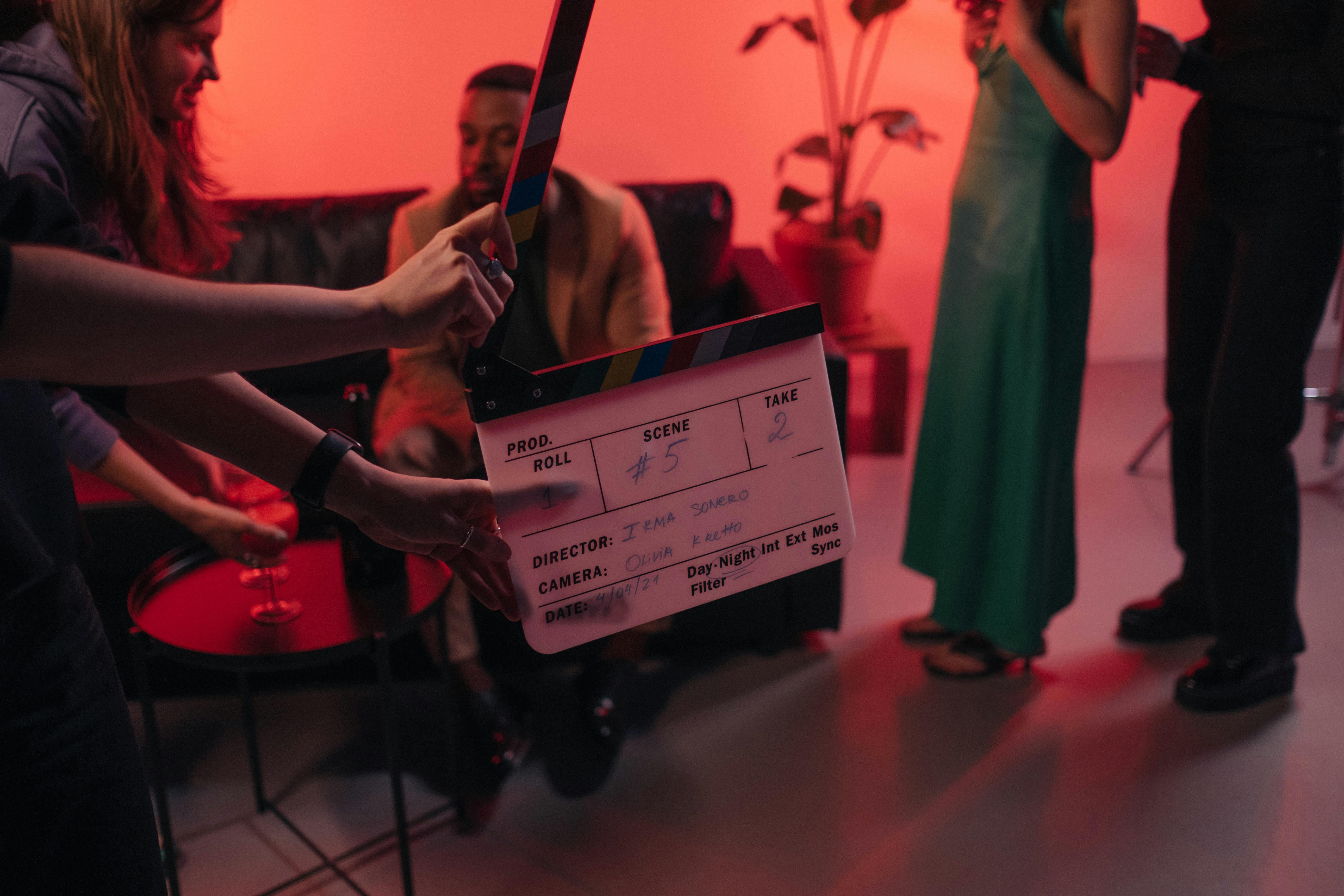 Hands hold a clapperboard for scene #5, take 2 on a film set with actors under dramatic red lighting.