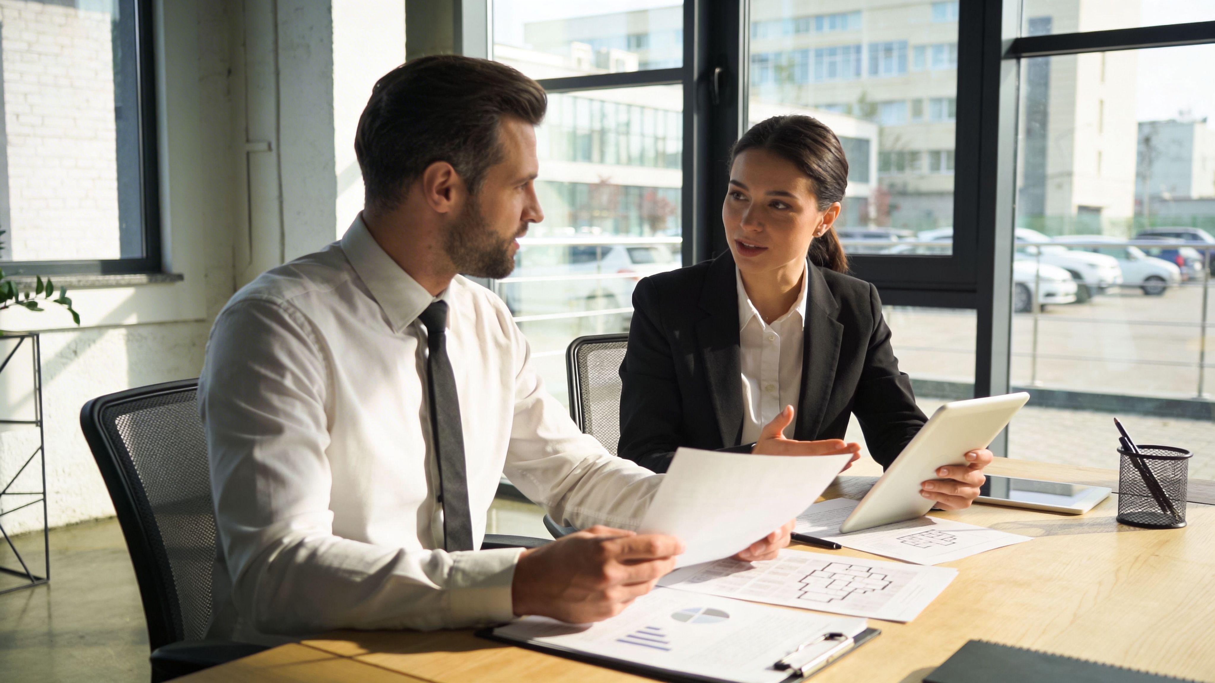 Two business professionals discussing reports and tablet data in a modern office setting