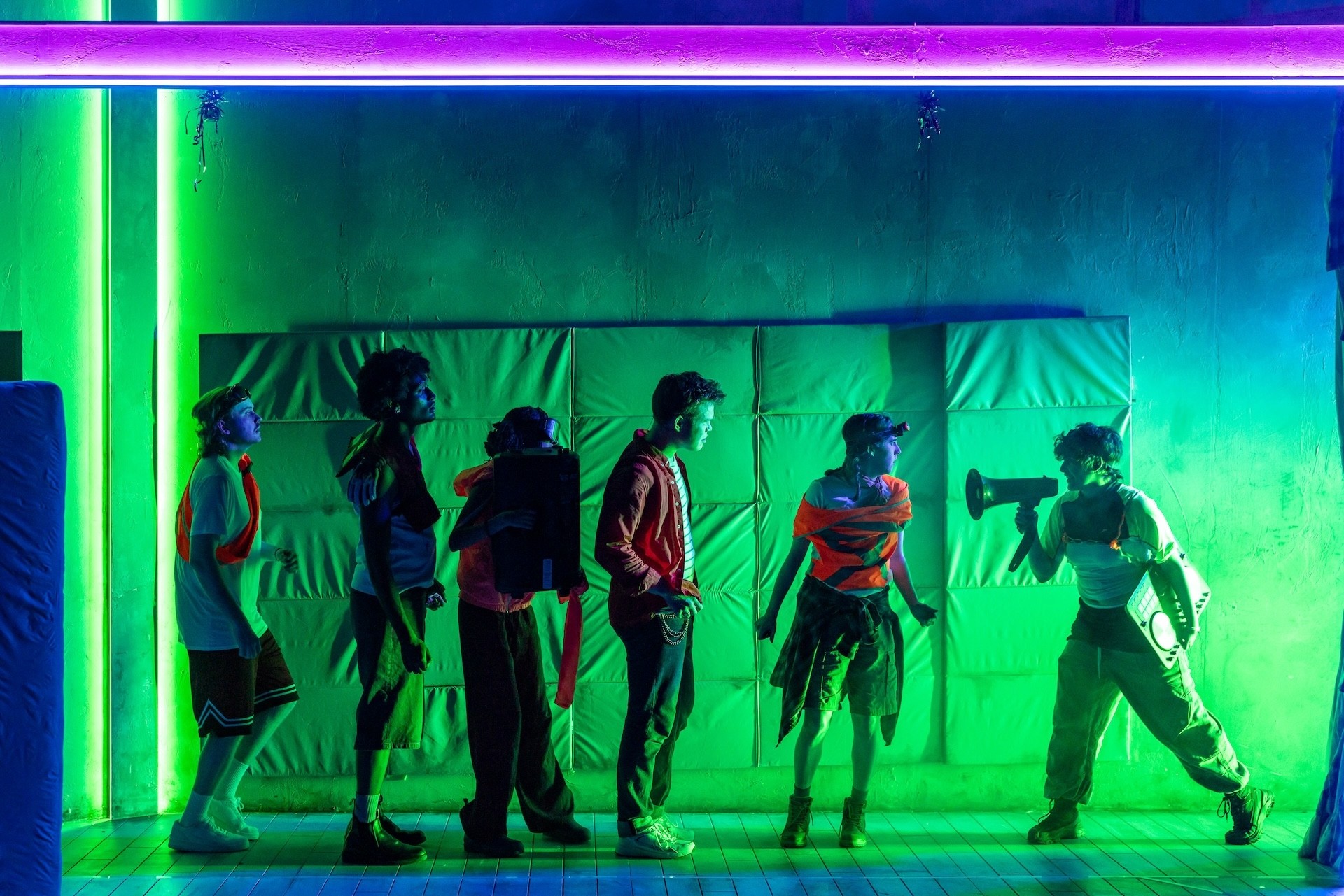 A line of young performers stands against a padded wall lit in vivid green and purple neon. One performer at the end raises a megaphone toward the group in a stylised, high-energy tableau.
