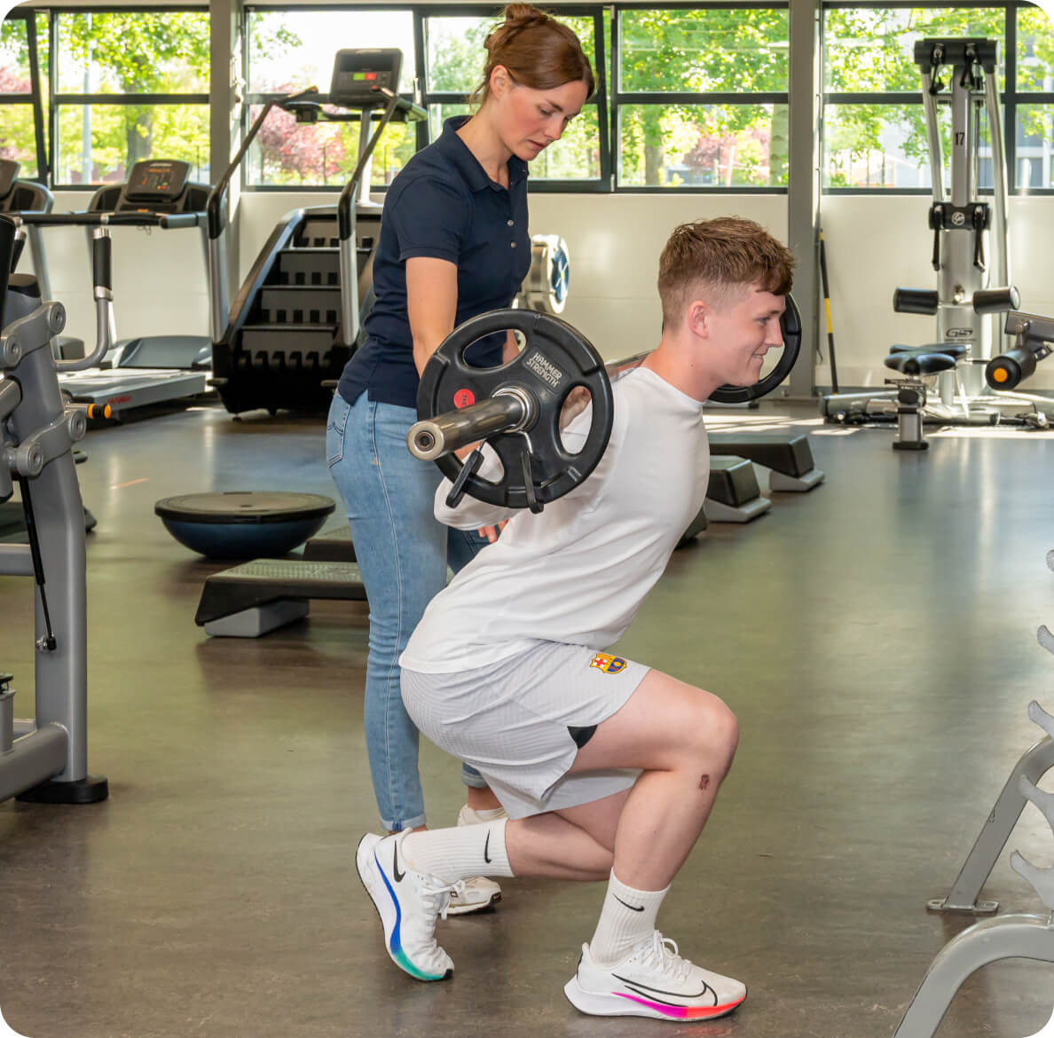 A person performs a barbell squat in a gym, assisted by a trainer, surrounded by modern fitness equipment and large windows letting in natural light.