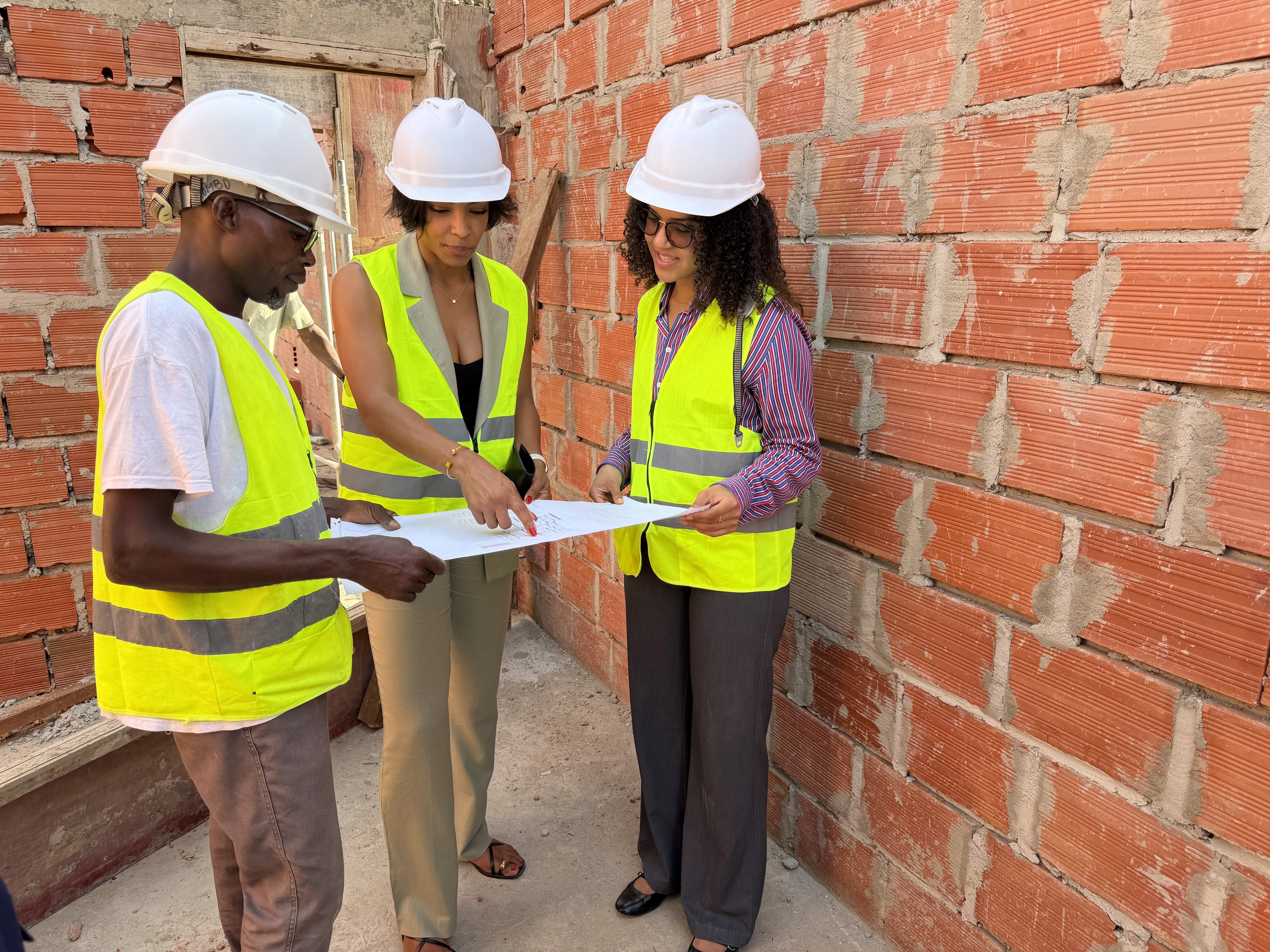 Three construction workers in safety vests and helmets review blueprints at a building site. The setting is sunlit, with a partially-built structure in the background.