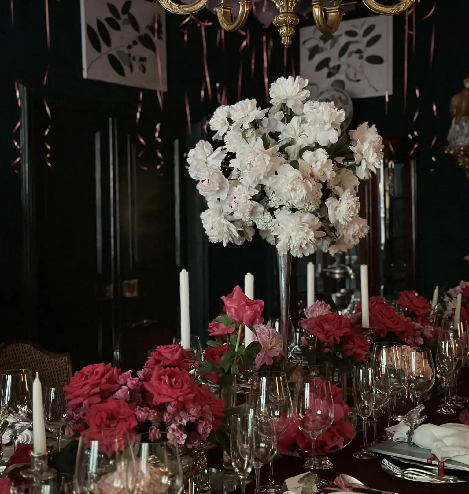 Mesa elegante decorada con centros florales en tonos blanco y fucsia, velas altas y copas de cristal bajo una lámpara de araña dorada, en un salón oscuro adornado con globos y cintas rosadas que cuelgan del techo.