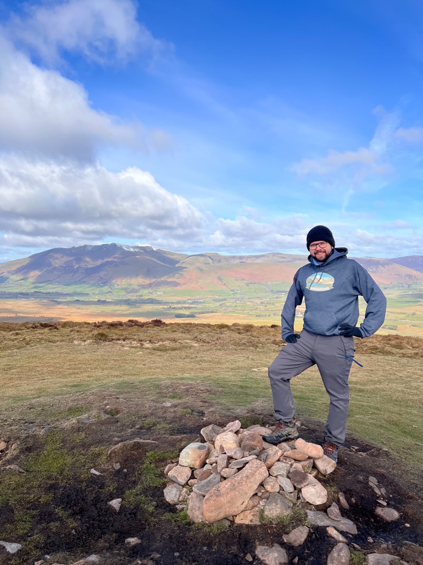 Martin stands with one foot on top of the stones at the summit of Great Mell Fell with his hands on his hips.