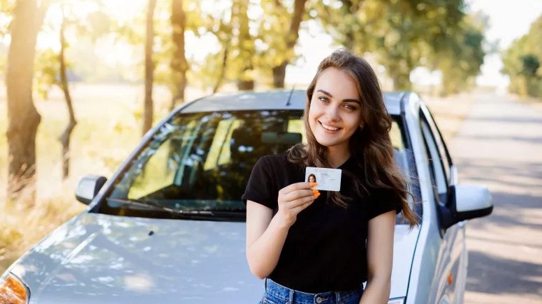 Młoda kobieta z uśmiechem trzymająca prawo jazdy, stojąca przy srebrnym aucie.