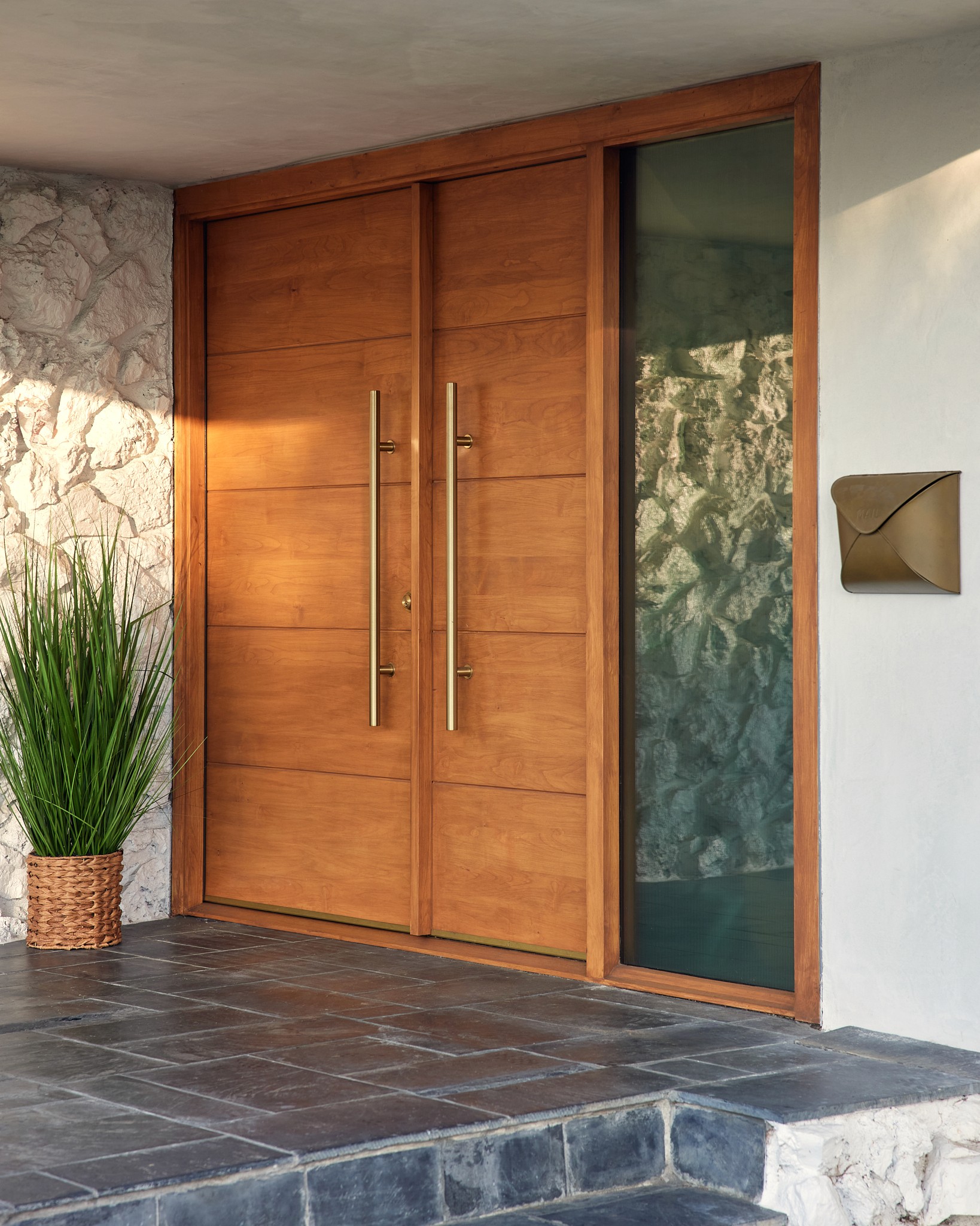 Modern exterior front door with warm wood, dark bricks, greenery, and white textured stucco statement wall by Jubilee Interiors in Los Angeles, California