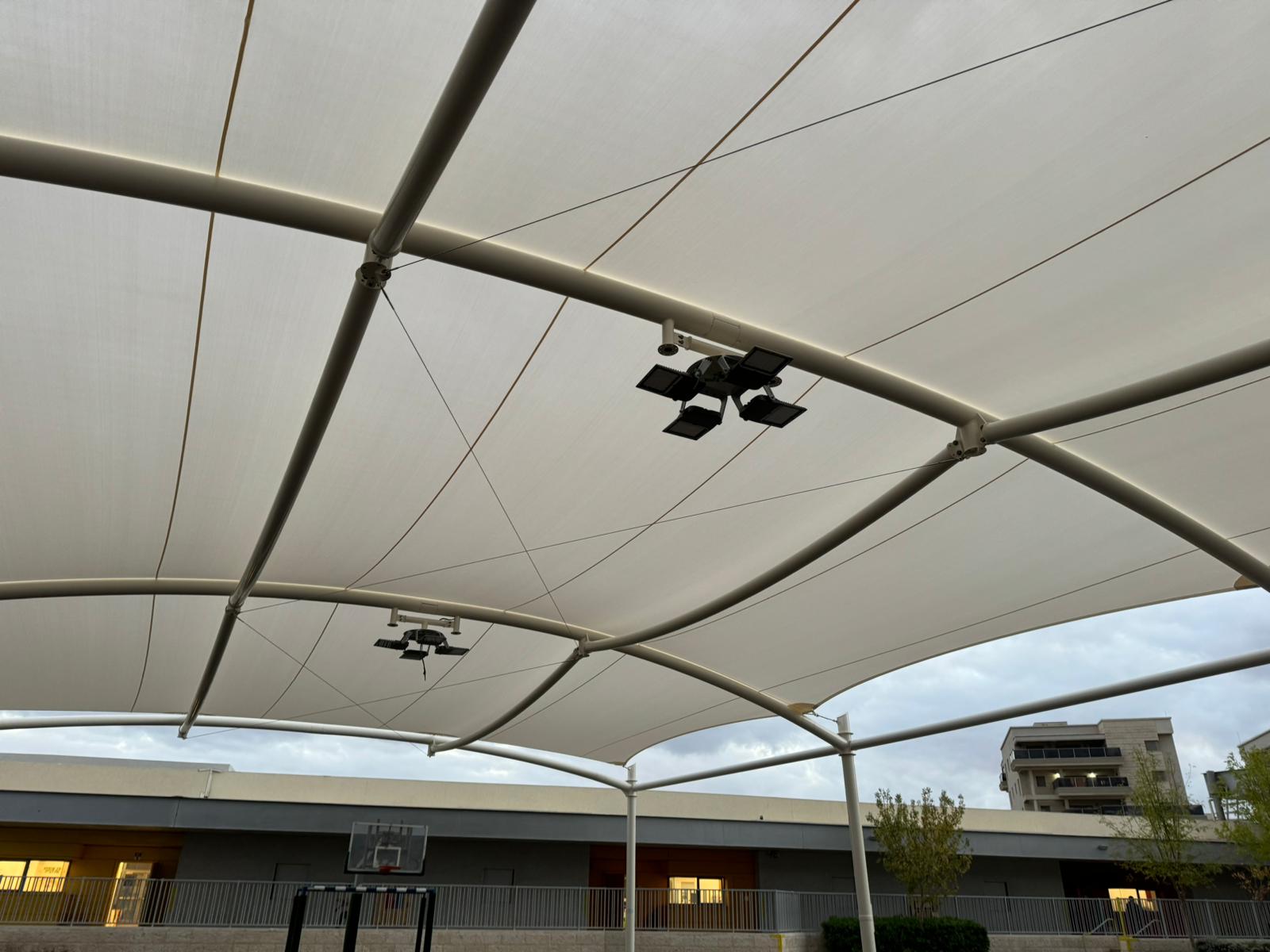 View under a large beige tensioned fabric roof with metal beams and floodlights above an outdoor basketball hoop.