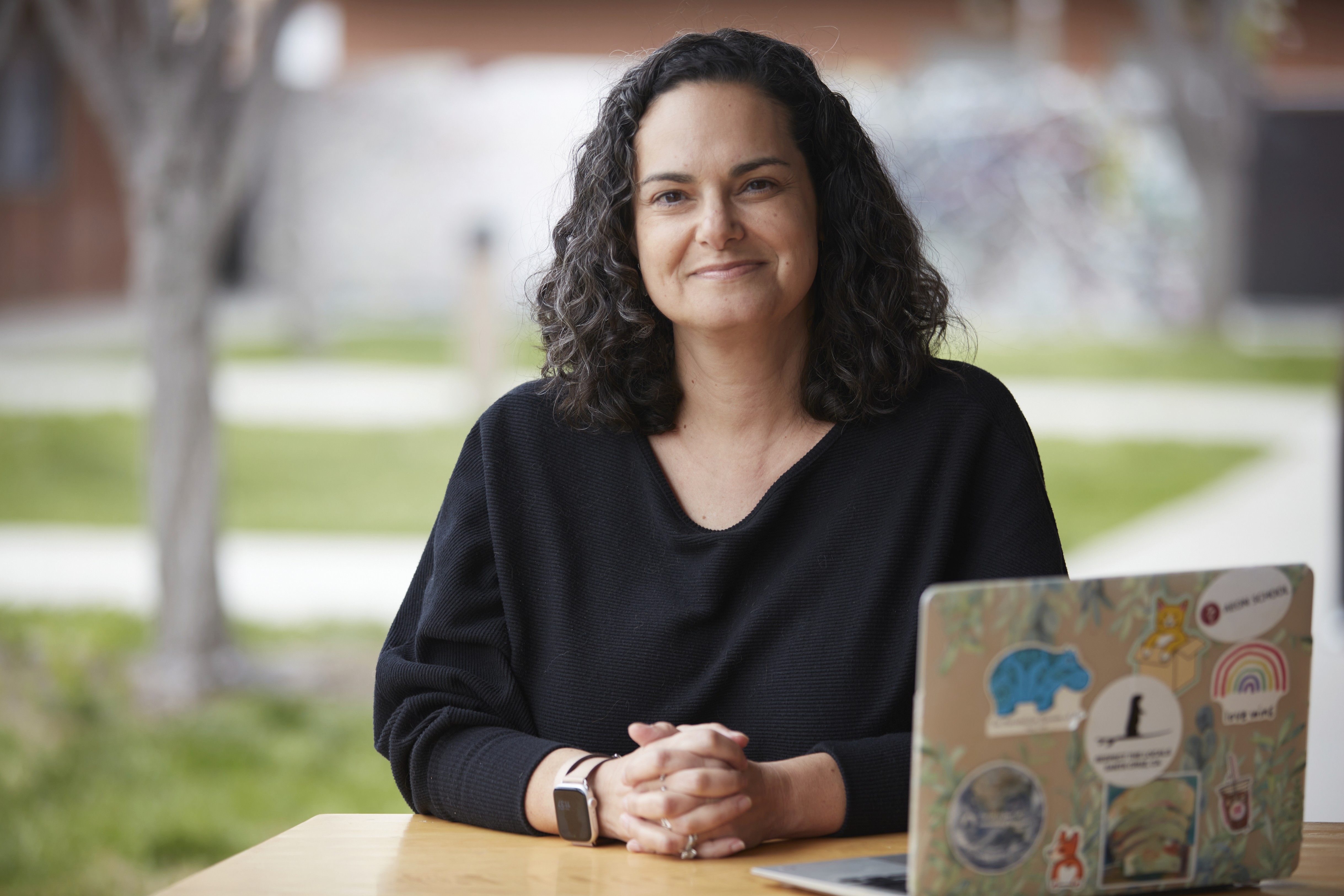  A young person with curly hair, wearing a grey sweatshirt, is seated in the driver's seat of a car, holding the steering wheel. An older man, wearing a striped shirt, is leaning down from outside the car, looking in through the open driver's window and smiling, while pointing toward the front of the car, seemingly giving driving instructions.
