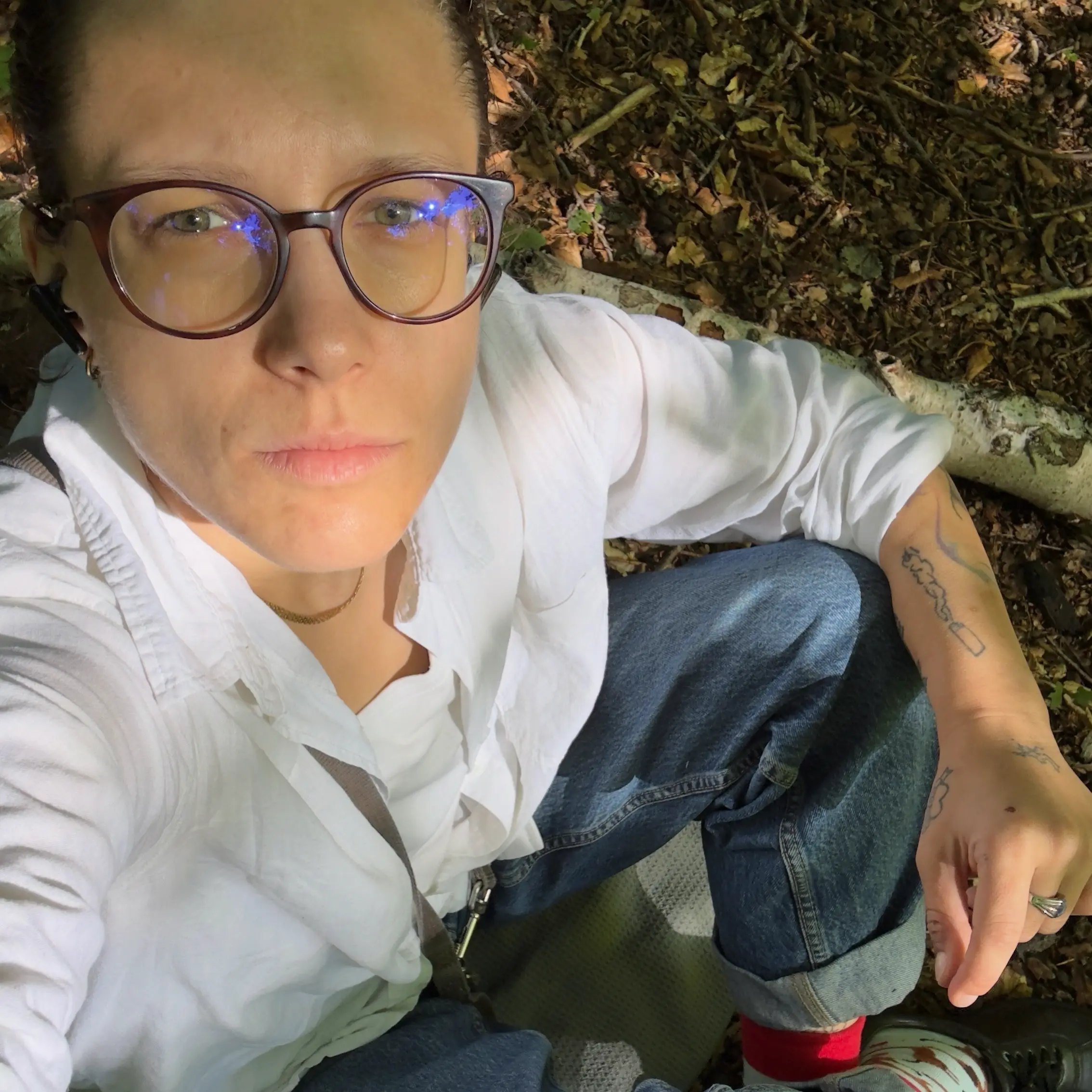 A woman with glasses sits outdoors in a forested area, wearing a white shirt, blue jeans, and red socks, and is surrounded by natural elements like leaves and branches.
