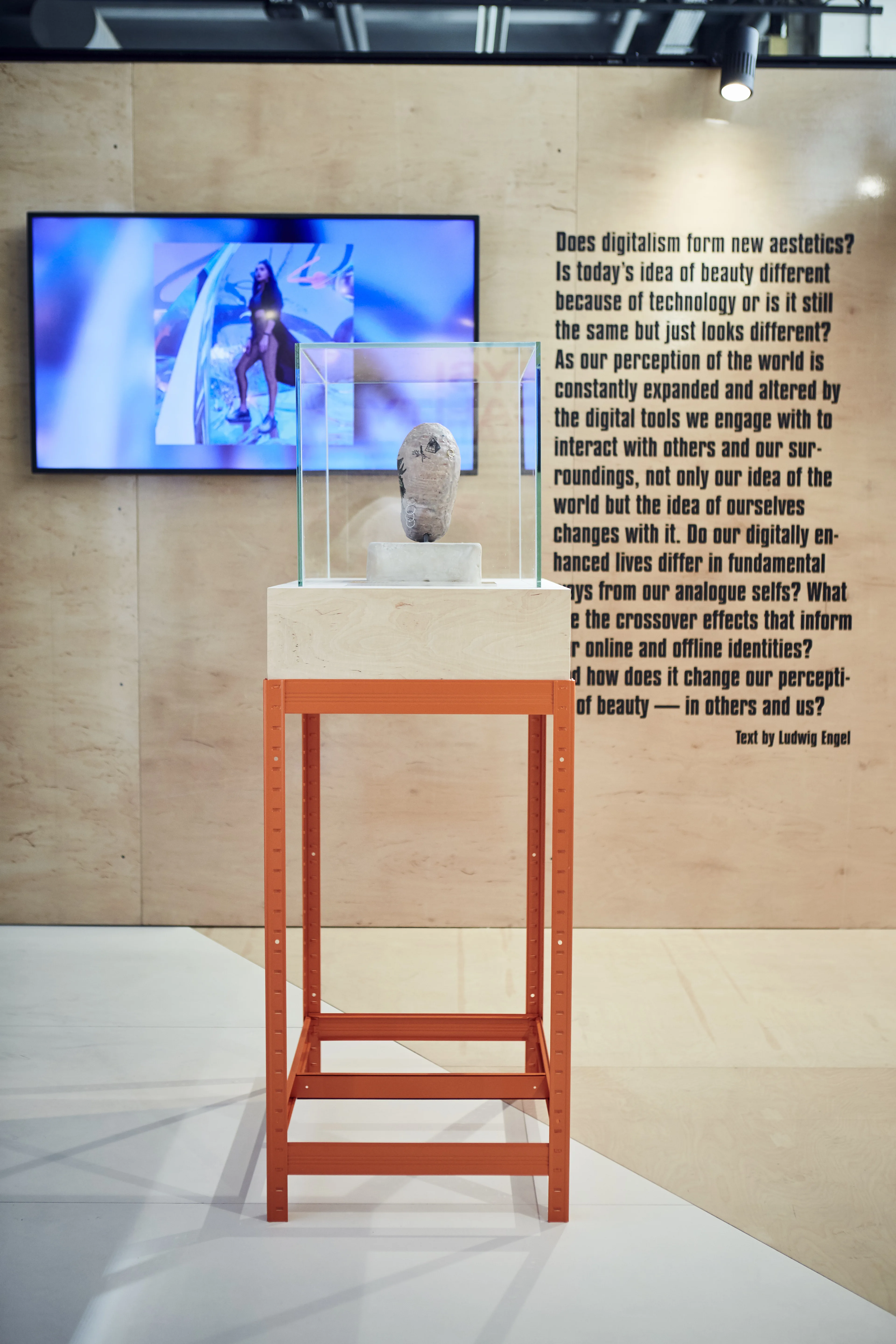 Sculptural exhibit in a glass case on an orange steel stand beside a screen and an essay on digital beauty at the Cosnova exhibition.