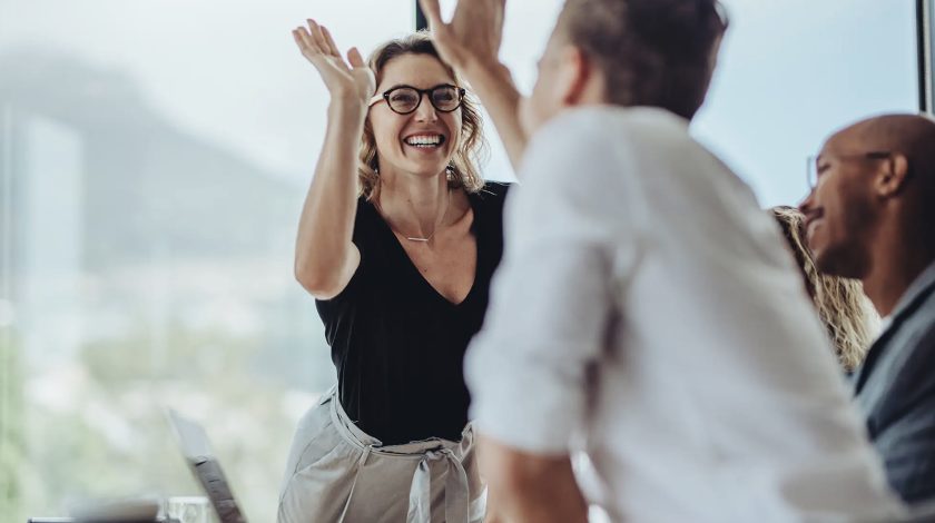 A smiling woman wearing glasses and a black top enthusiastically gives a high-five to a colleague during a daytime meeting in a bright, modern office.