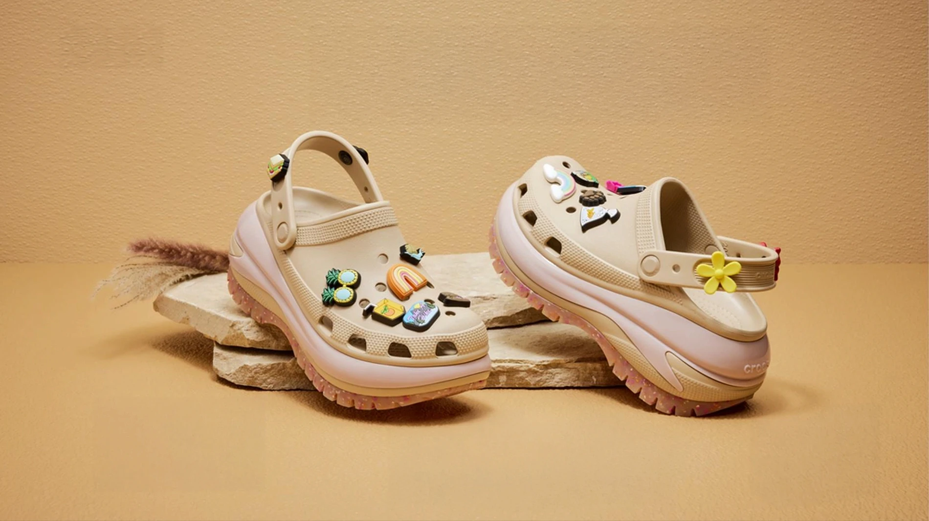 Crocs Classic Mega Crush clogs in cream and pink displayed on stone slabs, decorated with colourful Jibbitz charms including rainbow, flower and tropical designs, warm beige studio background