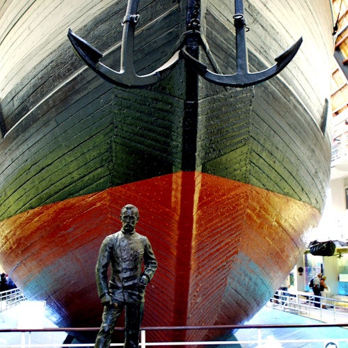 Крупный нос корабля с двумя якорями, выставленный в помещении над перилами. Статуя мужчины в морской форме стоит перед ним.