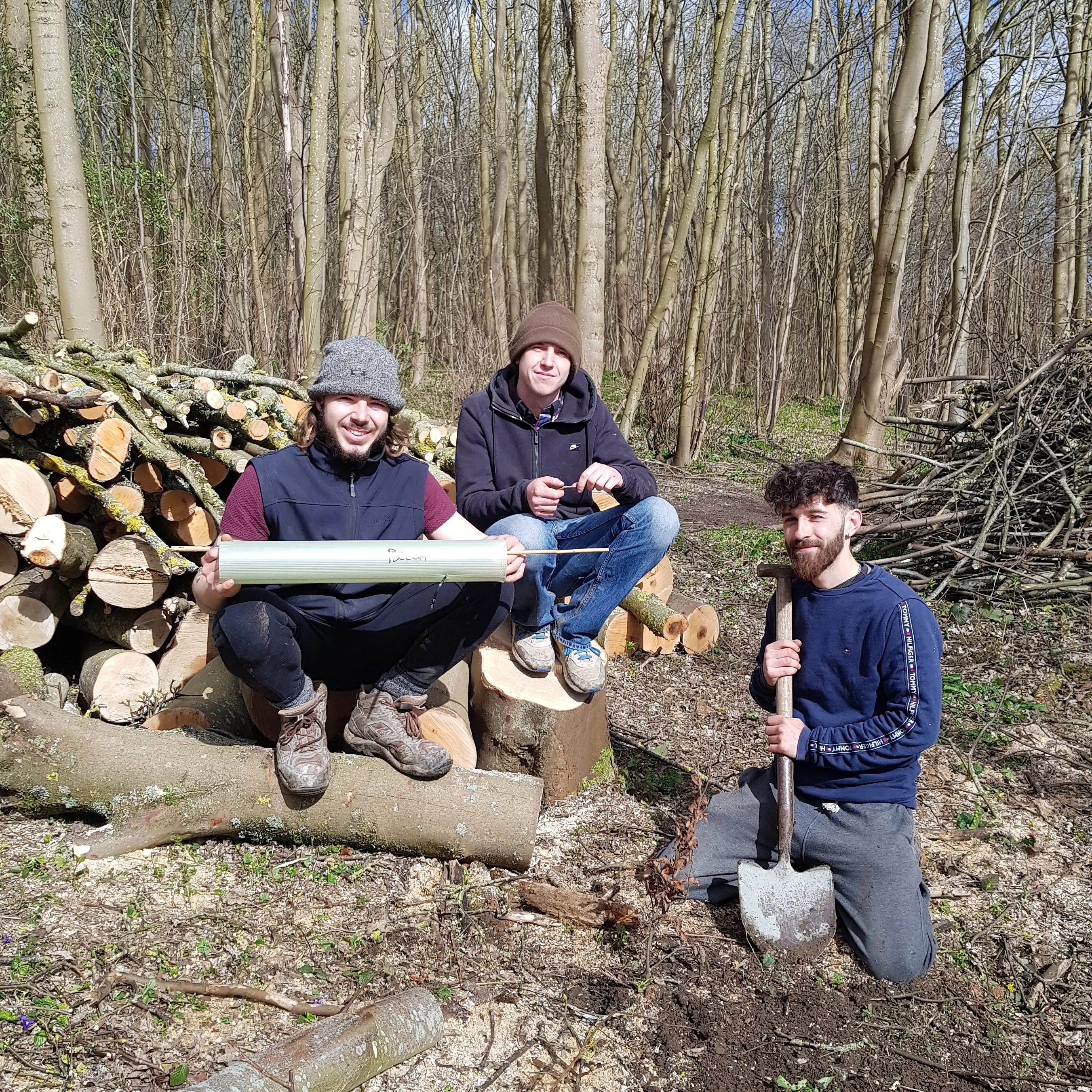 Three people in a forest, two sitting on logs and one crouching, surrounded by wood and nature.