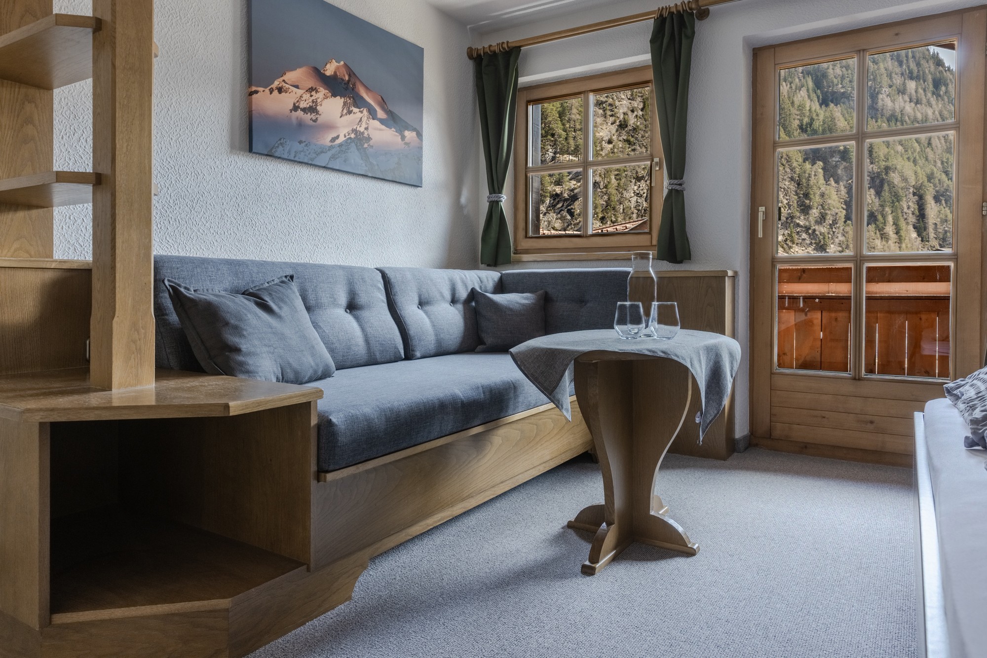 Gemütliches Wohnzimmer mit einem grau gepolsterten Sofa, einem Beistelltisch aus Holz und einem kleinen runden Tisch mit zwei leeren Gläsern vor der Kulisse großer Fenster mit malerischem Bergblick und einem Balkon, der von sanftem Tageslicht erhellt wird.