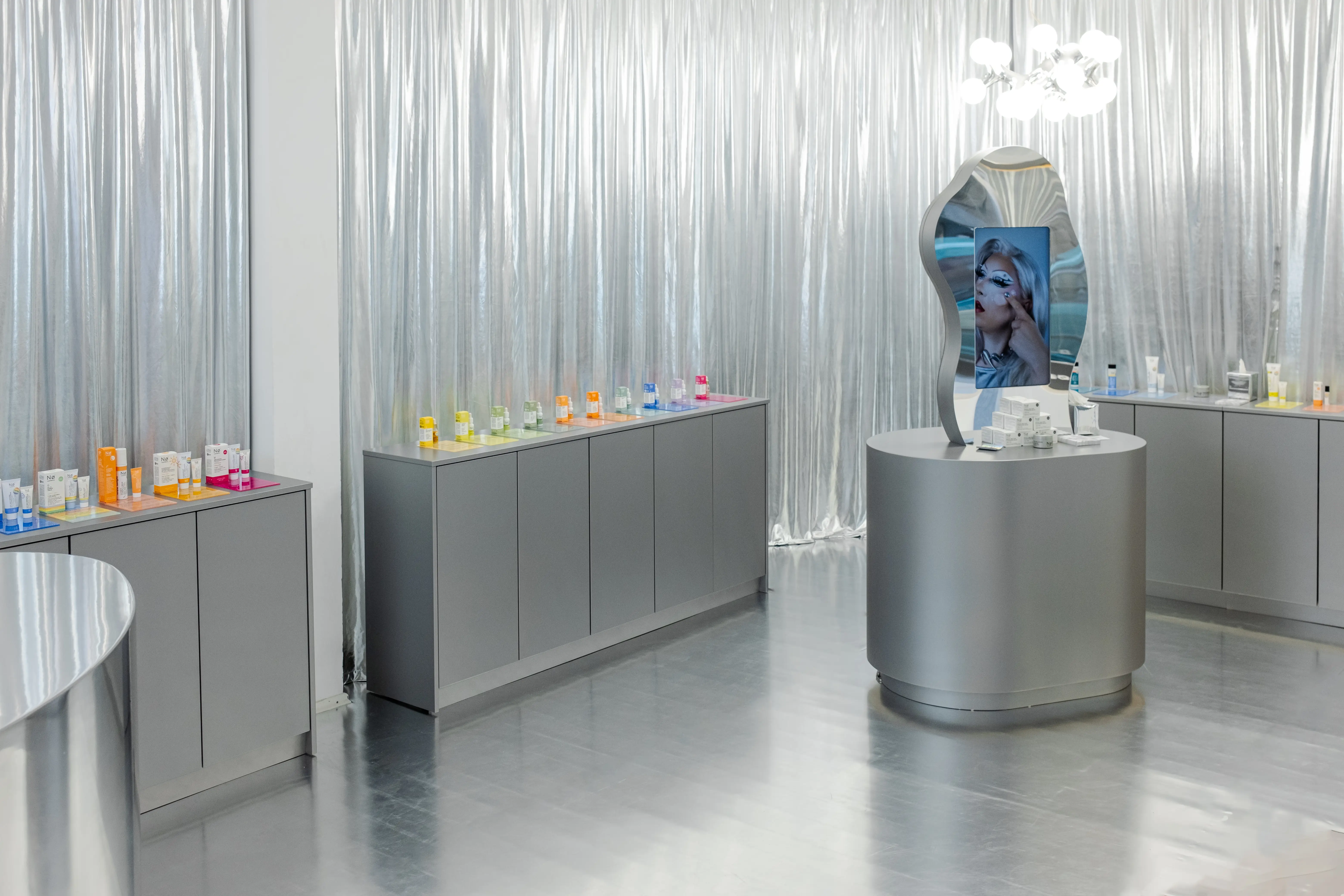 Silver showroom of NØ Cosmetics with metallic cabinets, colorful product rows and wavy mirror display.