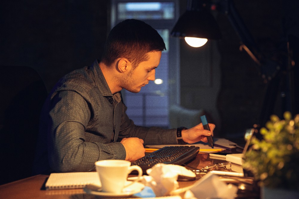 A man sitting at a table indoors, writing in a book. The room is dimly lit by a desk lamp. A coffee cup and saucer are placed on the table next to him.