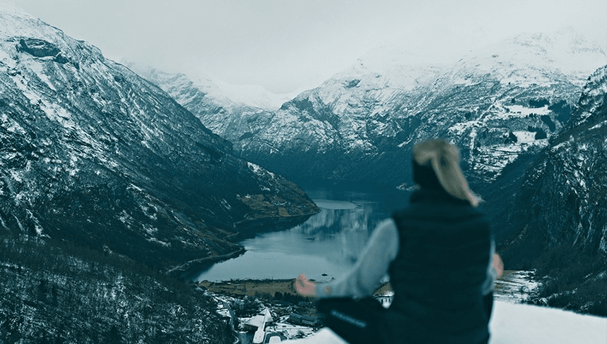 A person stands on a snowy mountain ledge, looking out over a stunning mountain landscape with snow-capped peaks.