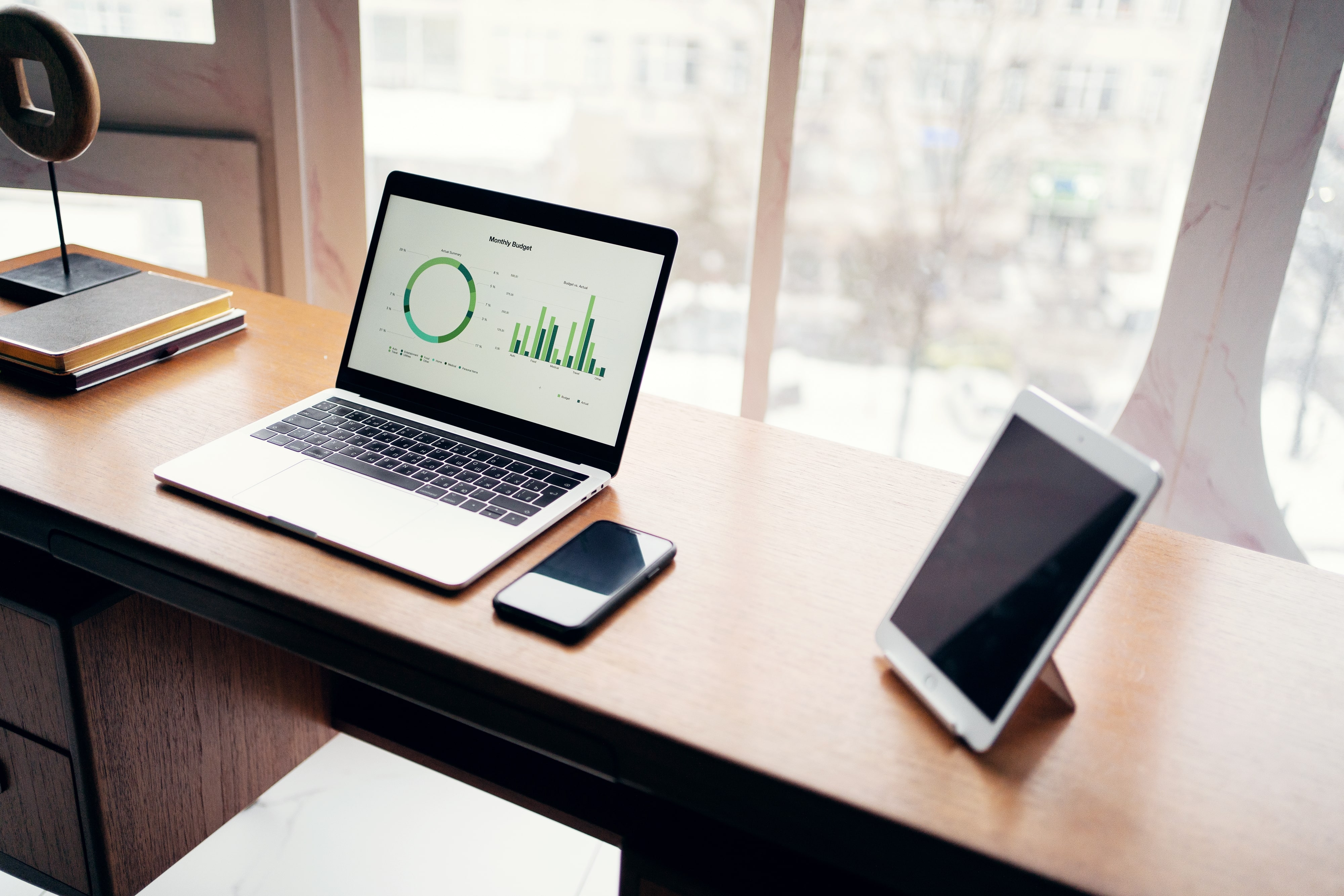 A modern home office workspace featuring a laptop on a wooden desk, displaying a "Monthly Budget" report with financial charts. Next to the laptop are a smartphone and a tablet, all set before a large, bright window, creating a productive remote work scene.