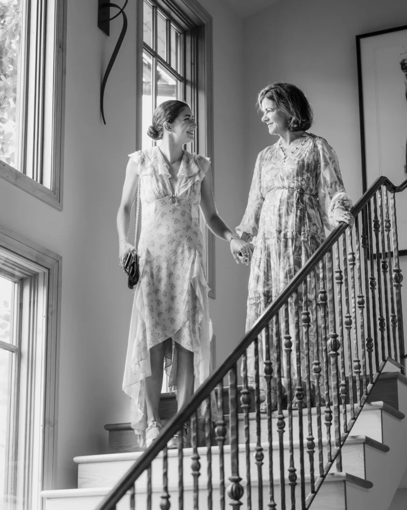 A black and white candid shot of two women in floral dresses holding hands while walking down an indoor staircase at a Chaparral Ranch Wedding.