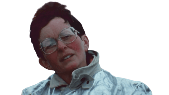 A rugged, close-up photograph of a woman wearing a heat-resistant silver suit and large glasses, standing near an active volcanic landscape with visible lava in the background. Her windblown hair and sunburned face suggest resilience and dedication, evoking the adventurous spirit of a volcanologist or field researcher studying extreme environments.