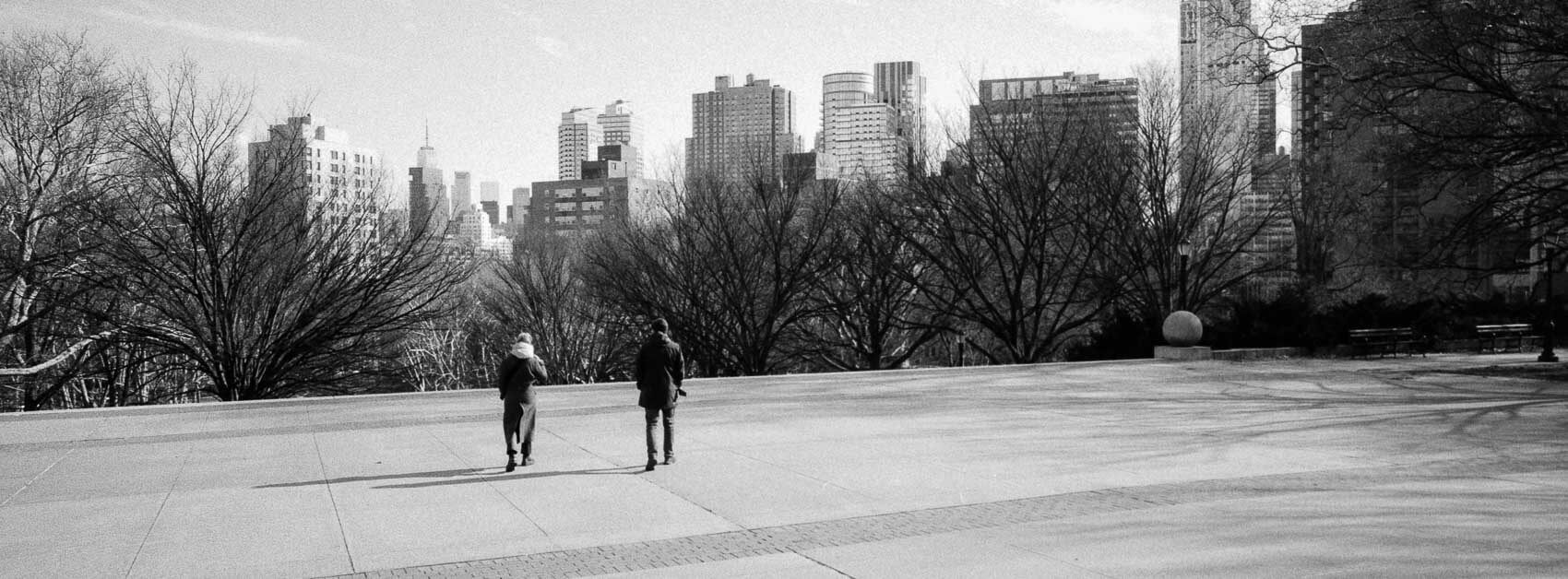 NYC pictures black and white a famous view, where people in the front walk downtime stairs and NYC skyline is visible in the background above the peaks of the tress in the park, panoramic Hasselblad XPan depth collection.