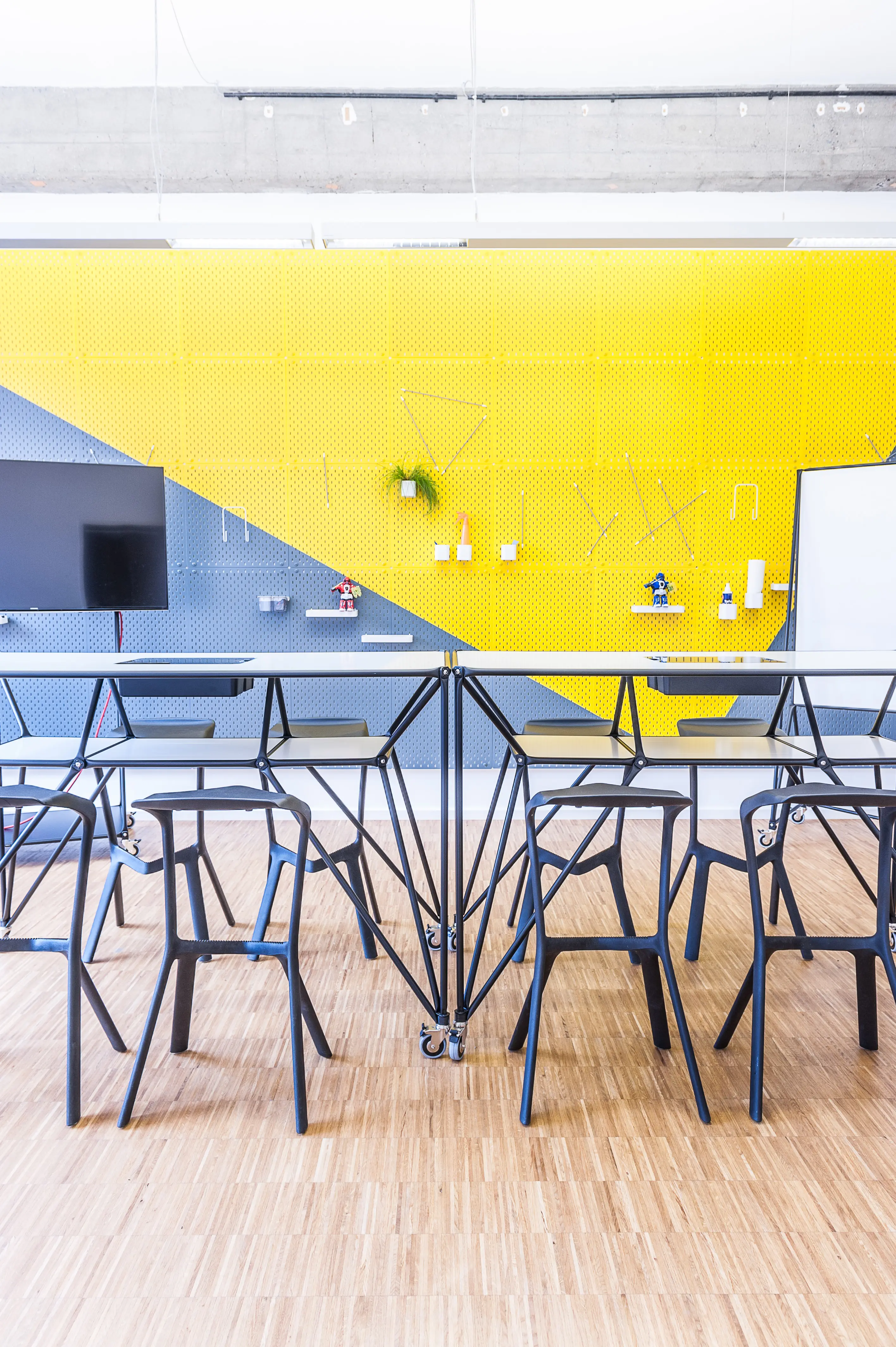Workshop area at BASILICOM with black Magis Spun-style chairs around mobile folding tables, a yellow-and-grey perforated pegboard wall with shelves and tools, and a wall-mounted screen.