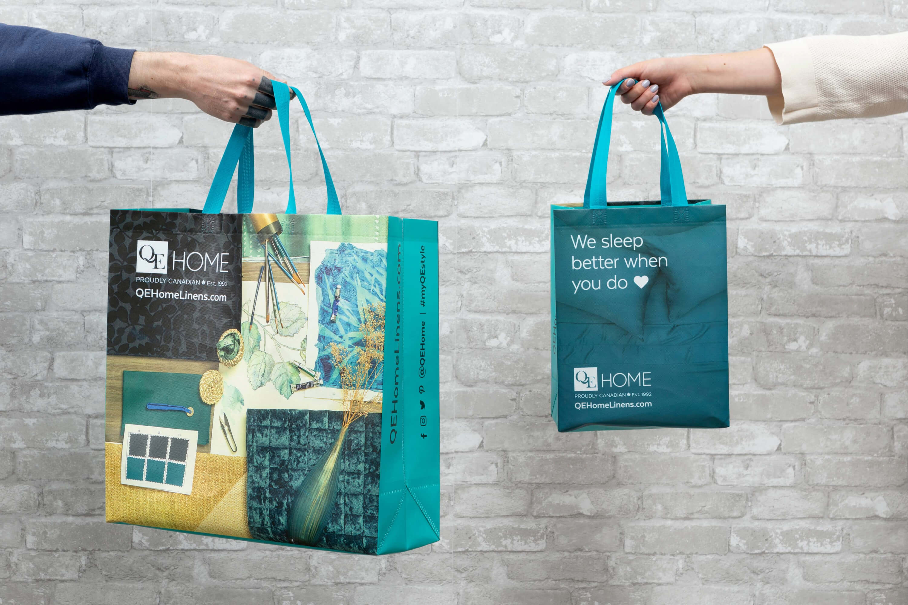 Two QE Home reusable shopping bags held up against a brick wall — one with a lifestyle print design and one with the 'We sleep better when you do' teal design