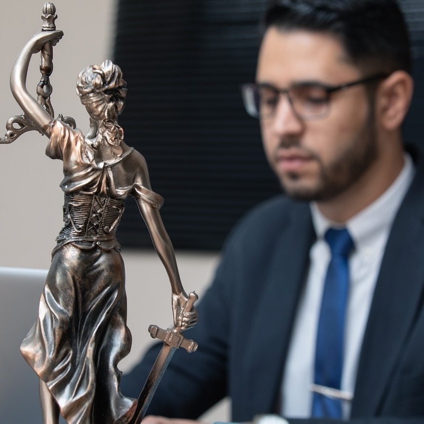 A small bronze statue of Lady Justice, and a man in formal wear in the blurred background.