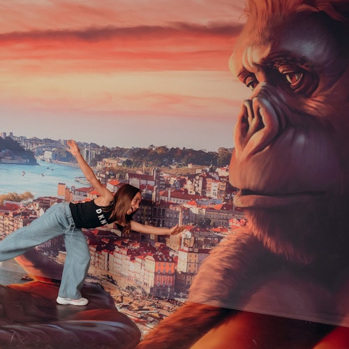 A woman poses playfully as if being held by a large King Kong figure against a scenic city and sunset backdrop.