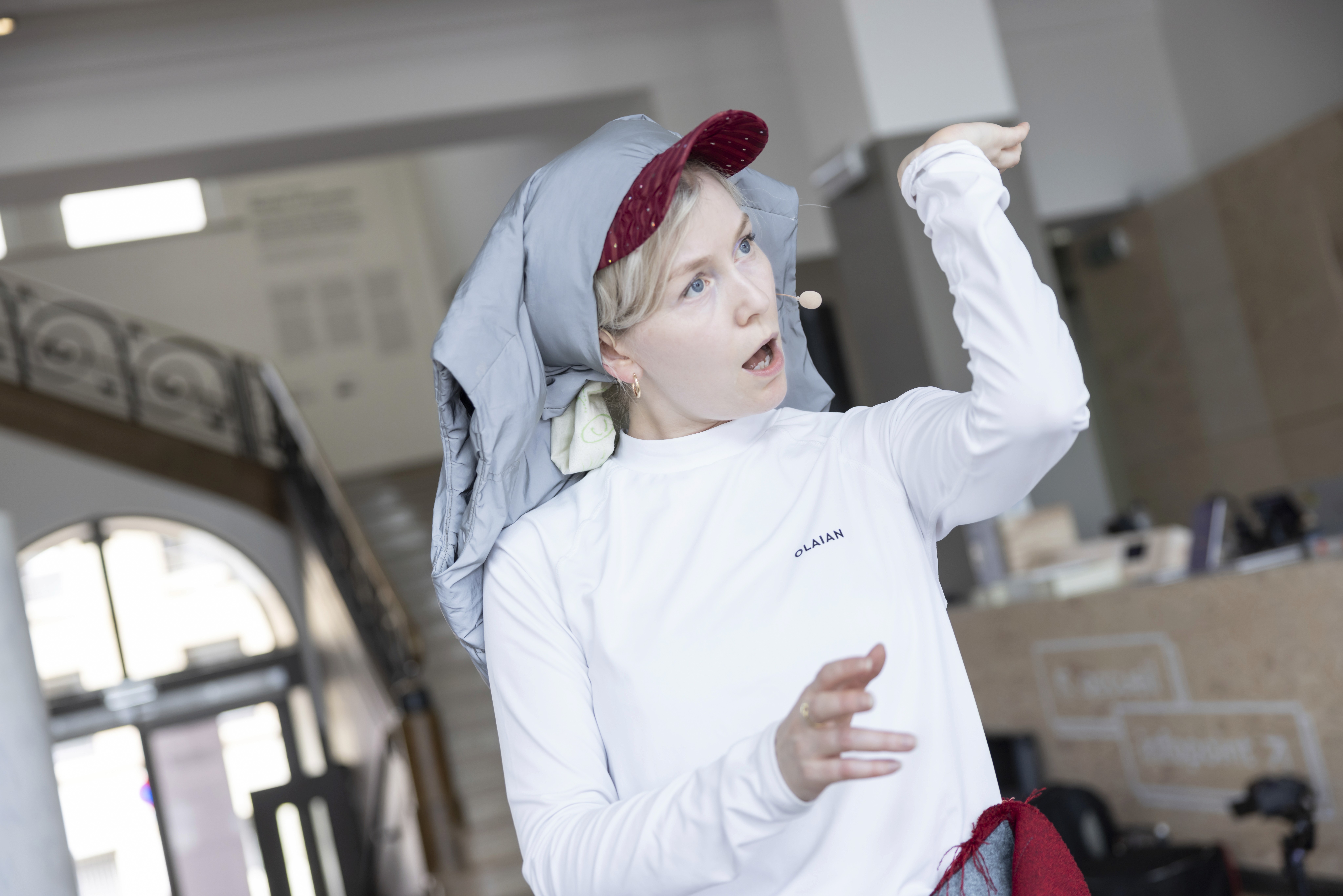 A still from Hyena Opera by Ásta Fanney Sigurðardóttir: the artist mid-performance, wearing a headset microphone and a white long-sleeve top, with a soft gray fabric draped under a red baseball cap on her head, one arm raised.