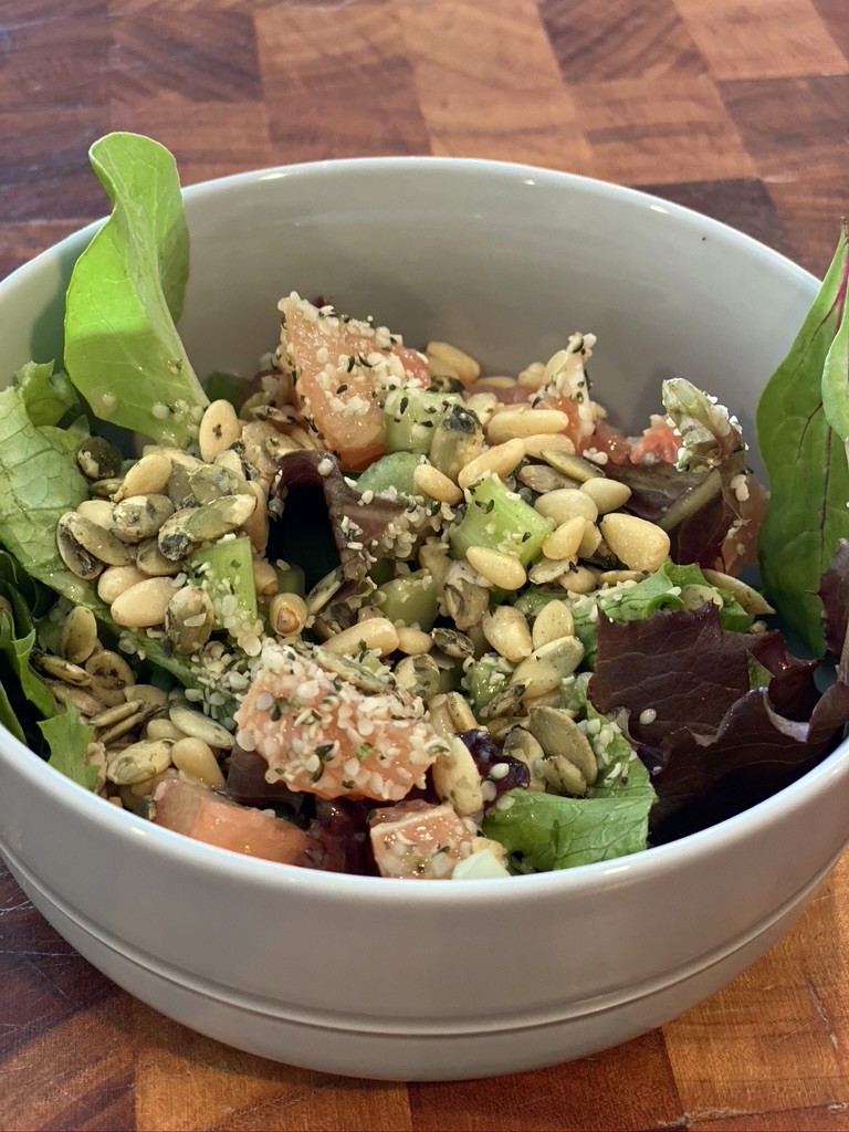A fresh salad in a white bowl features mixed greens, diced tomatoes, chopped cucumbers, and is topped with pine nuts, pumpkin seeds, and a sprinkle of hemp seeds, all placed on a wooden surface.