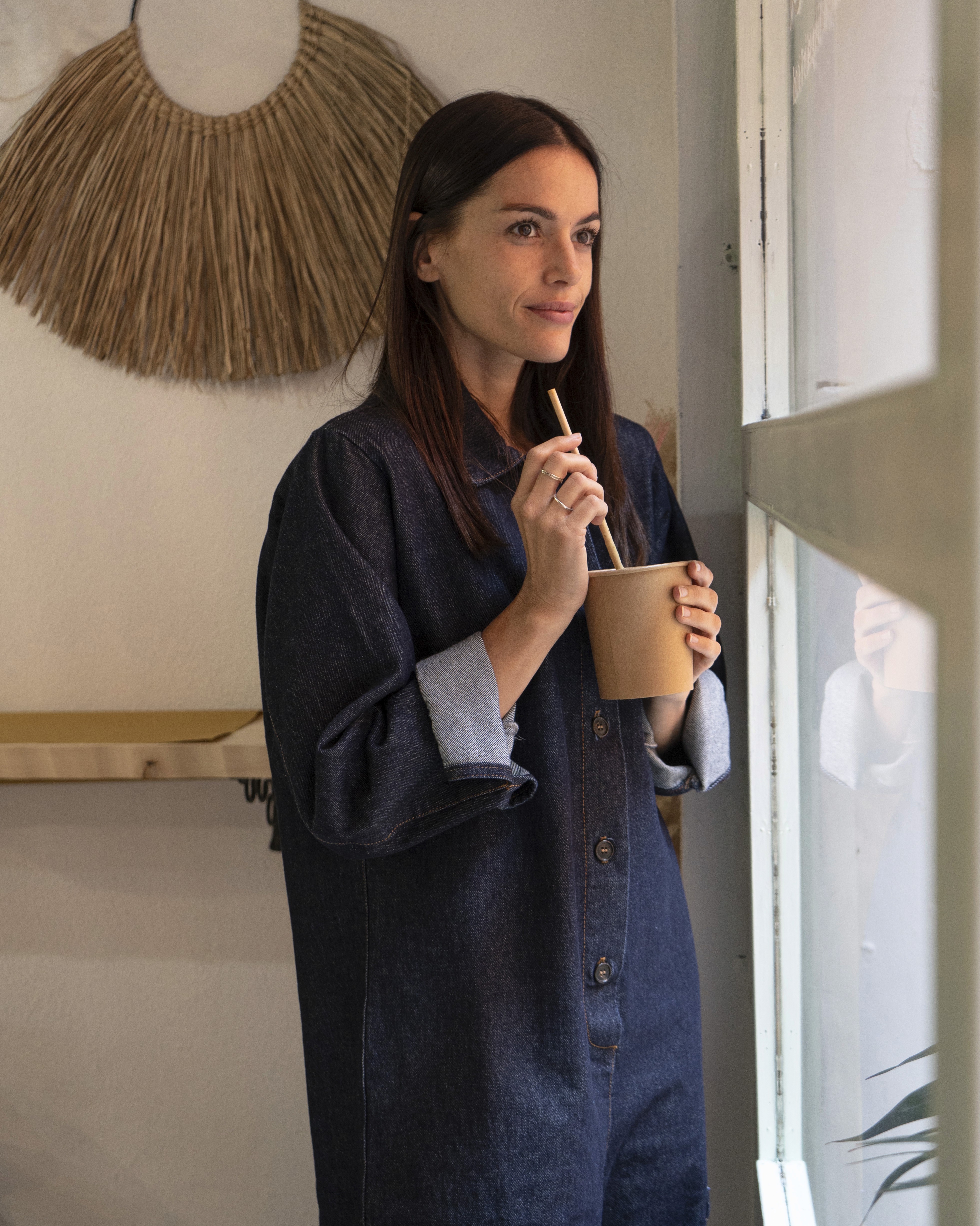 Photo de lookbook ou une femme portant une robe/chemise large en denim brut sourit et boit dans un gobelet en carton