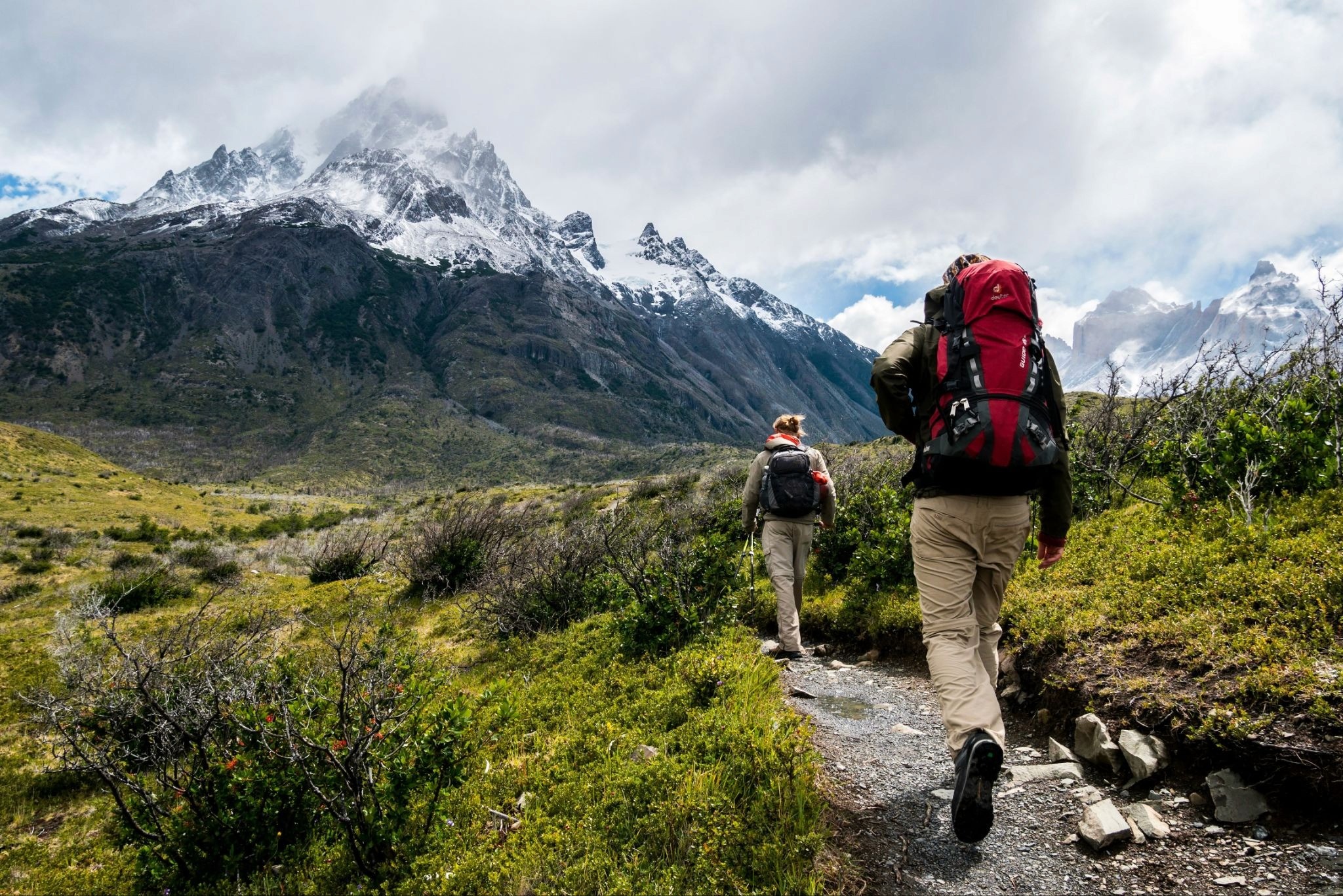 Two hikers trekking along a mountain trail beneath snowy peaks and cloudy skies.